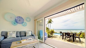 Un salon comprenant un canapé, une table basse et des objets décoratifs en verre. Il est doté d’une grande porte-fenêtre coulissante qui donne sur une terrasse offrant une vue sur l’océan et les palmiers.