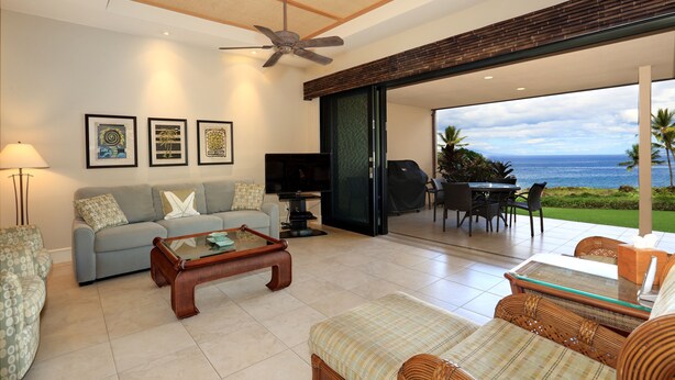 A spacious living room with a sofa, coffee table, and a television. There are framed pictures on the wall and a view of the ocean through the sliding glass door.