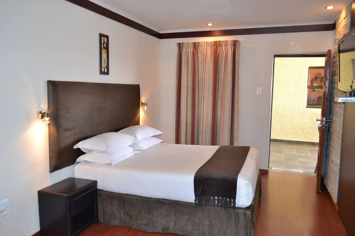Une chambre d’hôtel avec un grand lit, une table de chevet, une tête de lit, un rideau et une porte donnant sur une salle de bain.