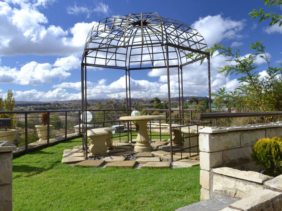 Um gazebo com estrutura metálica e base de pedra, cercado por um muro de pedra e uma área gramada com plantas em vasos.