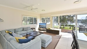 Ein geräumiges Wohnzimmer mit einem großen grauen Sofa, einem Flachbildfernseher und einem Balkon mit Blick auf das Wasser.