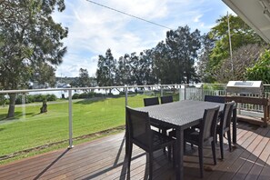A deck with a table and chairs, overlooking a grassy area and a body of water with boats. - Wangi Lakehouse Cottage  Lake Macquarie (Wangi Wangi)