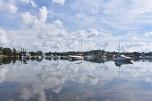 Ein Yachthafen mit mehreren vertäuten Booten, ruhigem Wasser, das Boote und Himmel reflektiert, und Industriegebäuden im Hintergrund.