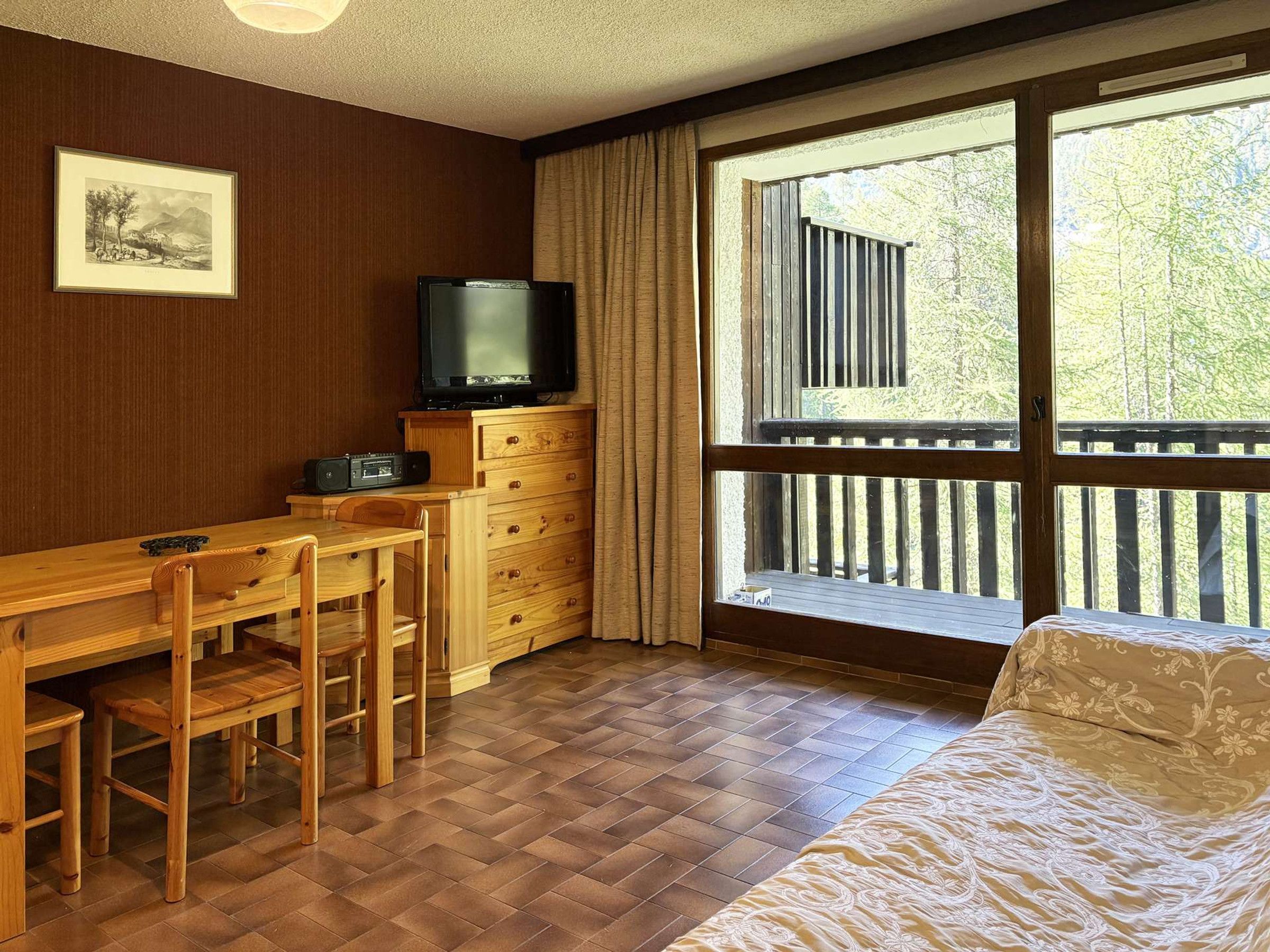 A room with a wooden desk, a television, a bed, and a window with a view of trees.