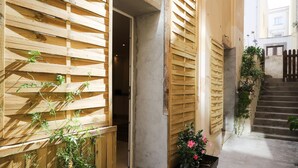 Un estrecho patio con contraventanas de madera, una planta en maceta y una escalera que lleva a un nivel superior.