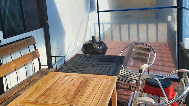 Un espacio de descanso al aire libre con techo, que incluye una mesa de madera, sillas de metal, una parrilla y vistas a un edificio a través de un panel transparente.