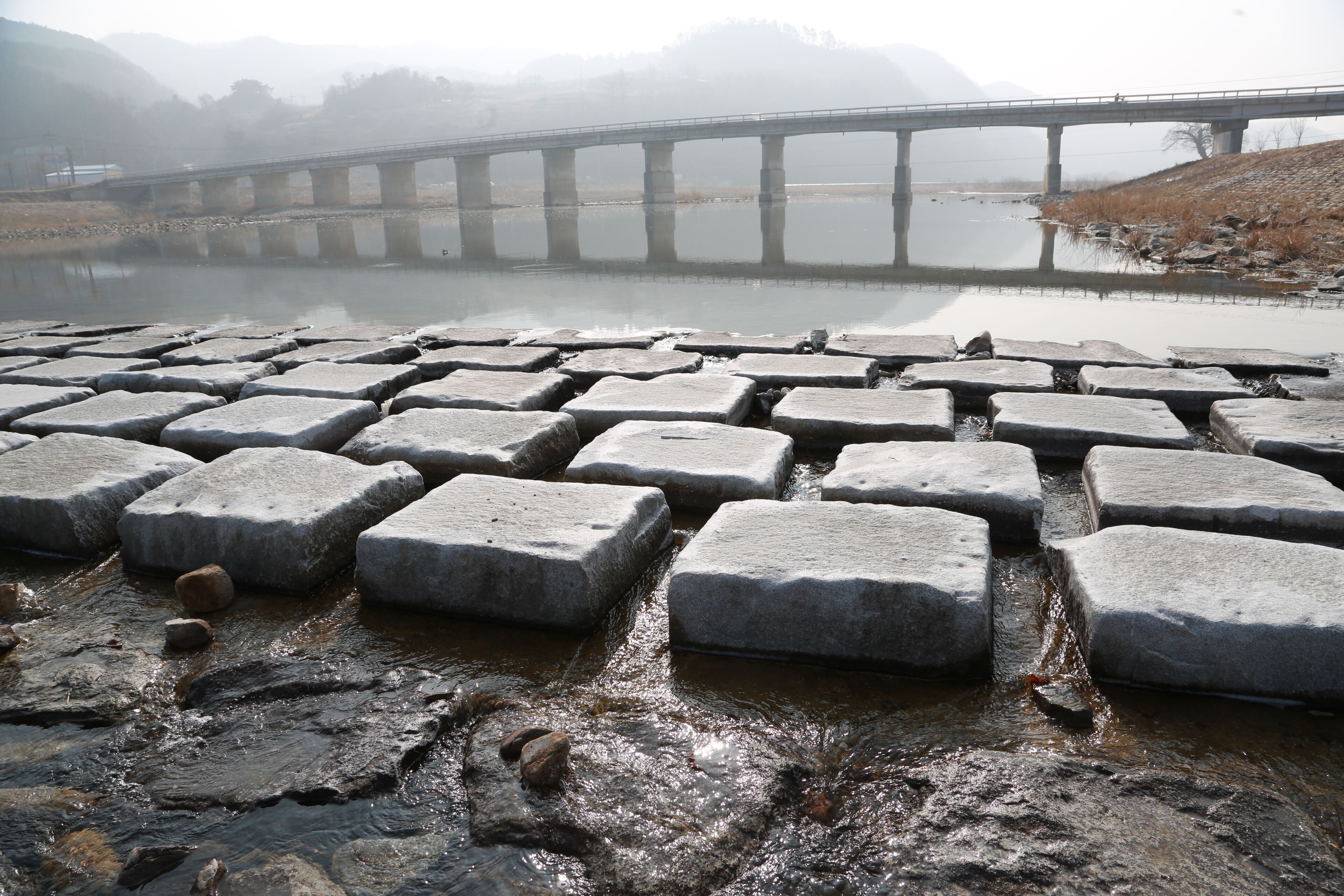 A stone bridge over a calm river with a misty background.
