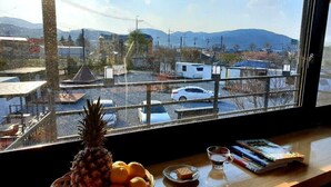 Vista desde una ventana sobre un estacionamiento con coches y edificios, una piña, naranjas, un vaso de agua y revistas sobre una mesa.