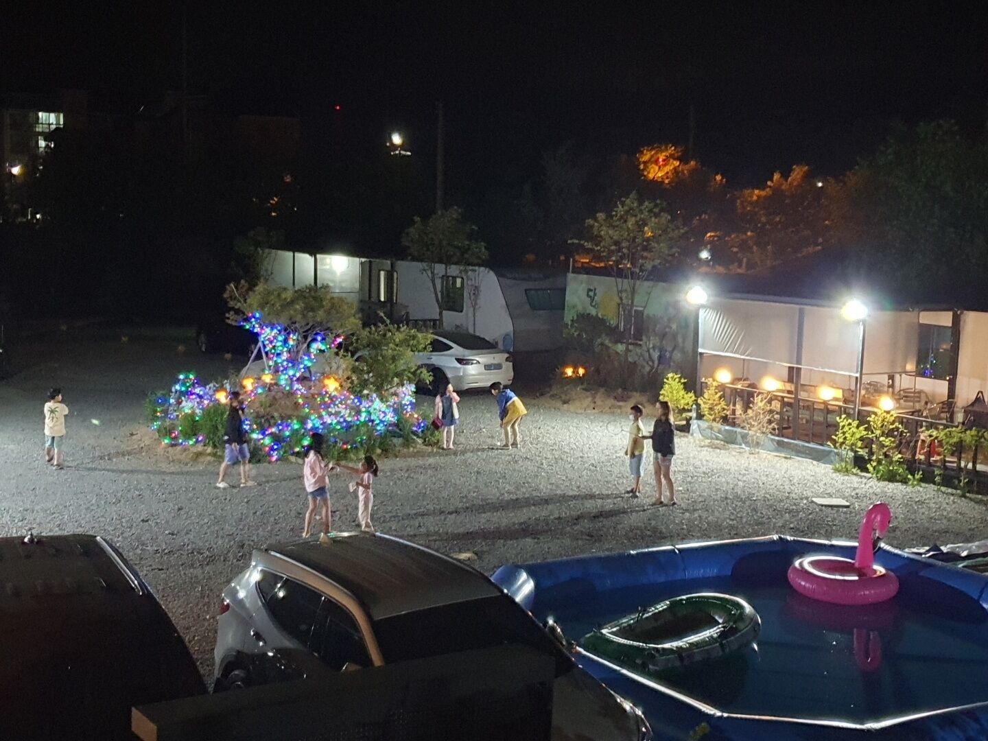 Un espace extérieur aménagé pour la nuit, avec un éclairage décoratif, une piscine agrémentée d’un flotteur rose et une voiture blanche garée à proximité.