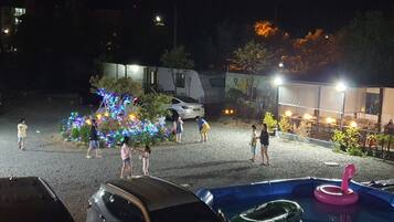Un espace extérieur aménagé pour la nuit, avec un éclairage décoratif, une piscine agrémentée d’un flotteur rose et une voiture blanche garée à proximité.