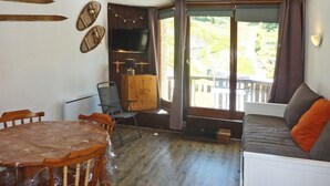 A room with a wooden dining table, chairs, a sofa, a TV, and a view of greenery outside. - Renovated studio, ski-in/ski-out, south-facing balcony (Les Orres)