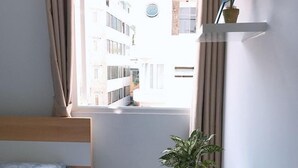 A small, bright room with a single bed, a round table with a plant, and a window overlooking a building. - Saigon House 18Bis (Ho Chi Minh City)