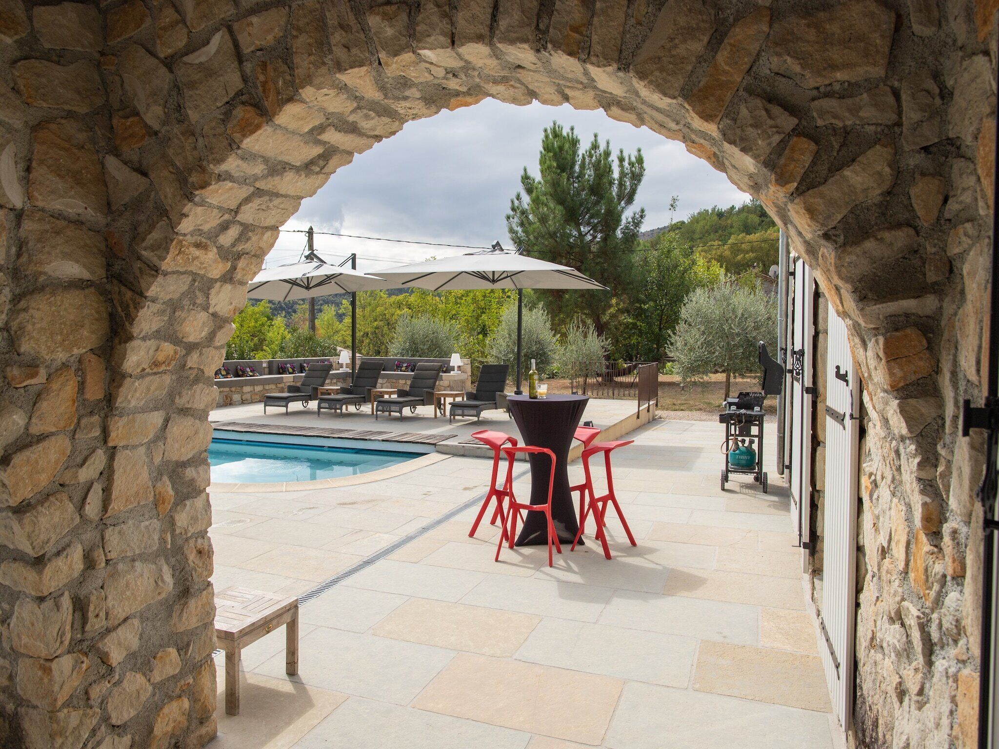 Un arc en pierre mène à un espace piscine agrémenté de chaises longues, de parasols et d’une table avec des chaises rouges.