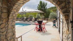 Ein steinerner Bogen führt zu einem Poolbereich mit Liegestühlen, Sonnenschirmen und einem Tisch mit roten Stühlen.