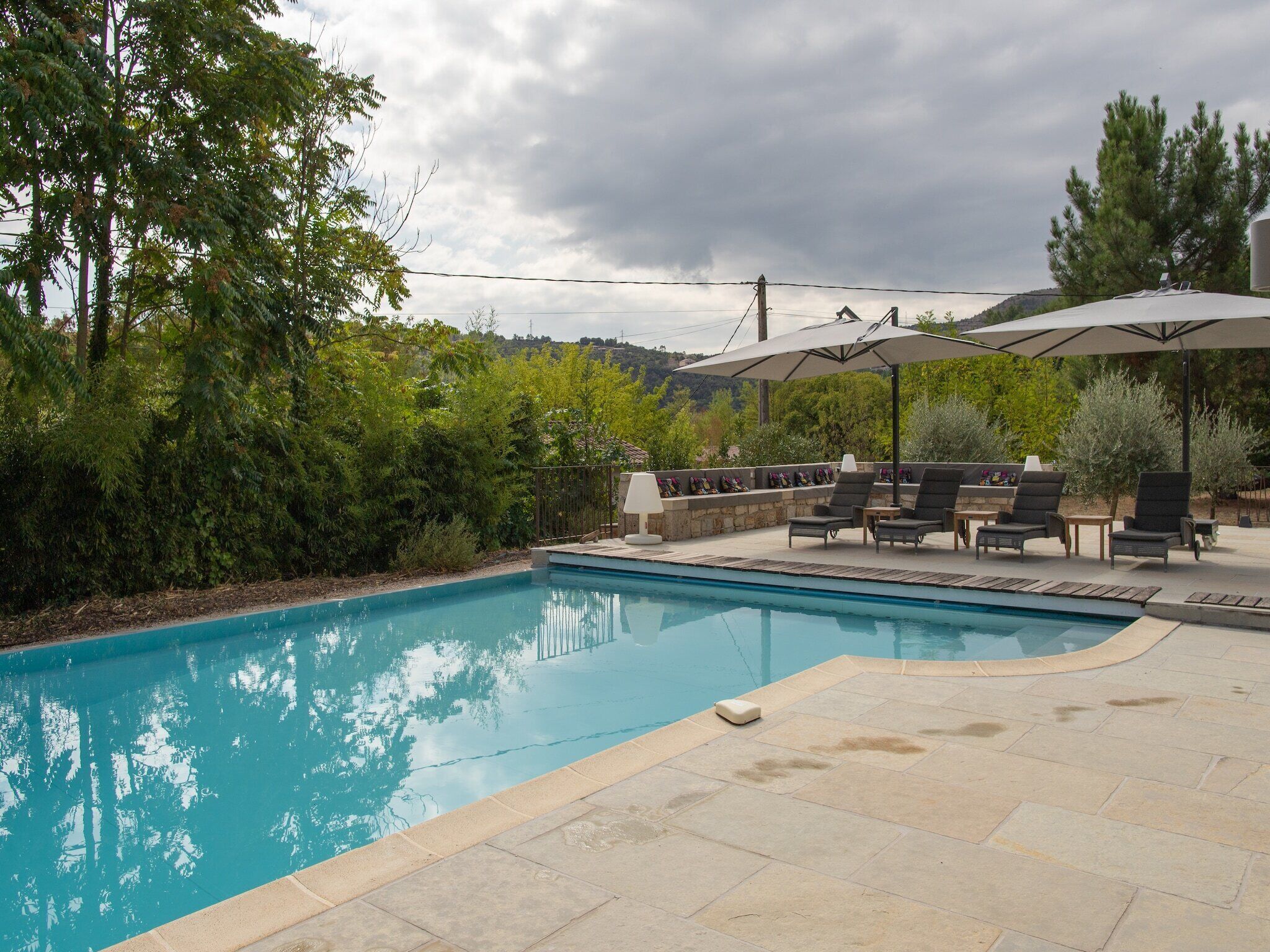 A swimming pool with lounge chairs and umbrellas, surrounded by greenery and a stone patio.