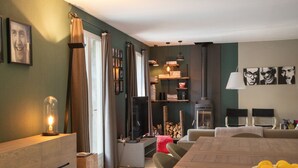 Ein modernes Wohnzimmer mit einem Holzesstisch, einer Couch und einem Fernseher. Vorhänge, eine Lampe und ein Bücherregal mit diversen Gegenständen sind ebenfalls zu sehen.
