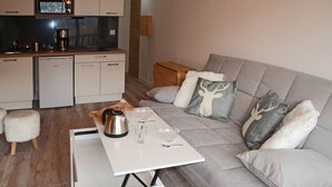 A modern living room with a grey sofa, a small white table, and a kitchen area in the background. - Newly Renovated Studio for 2 in Central Location with Direct Slope Access, Balcony & Stunning Views (Les Orres)