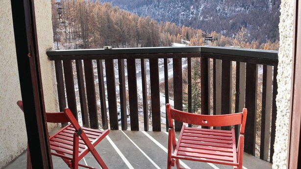 A balcony with two red chairs, a view of snow-covered mountains, and a parking lot in the background. - Newly Renovated Studio for 2 in Central Location with Direct Slope Access, Balcony & Stunning Views (Les Orres)