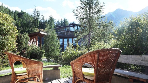 Zwei Korbstühle mit Kissen auf einer Holzdielenveranda mit Blick auf eine Berglandschaft, im Hintergrund ein modernes Gebäude.