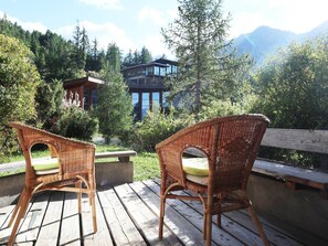 Deux fauteuils en osier, dotés de coussins, sont placés sur une terrasse en bois qui offre une vue sur un paysage montagneux, avec un bâtiment moderne en arrière-plan.