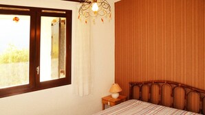 Ein Schlafzimmer mit Bett, Nachttisch, Lampe, einem Fenster mit Vorhängen und Wandjalousien.