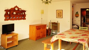 Ein Zimmer mit einem HolztV-Schrank, einem Bett mit Blumenmuster-Bettdecke, einer Holzkommode, einem Regal mit Küchenutensilien und einer Wandpflanze.