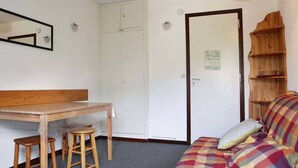 Una habitación con una mesa de madera, dos taburetes, un sofá con una manta escocesa, una estantería de madera y un armario blanco.