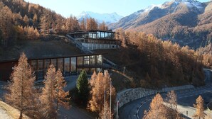 A mountain lodge with a glass facade, surrounded by autumn trees and a winding road. - Lovely studio for 4, ski-in/ski-out, wifi, pets allowed (Les Orres)