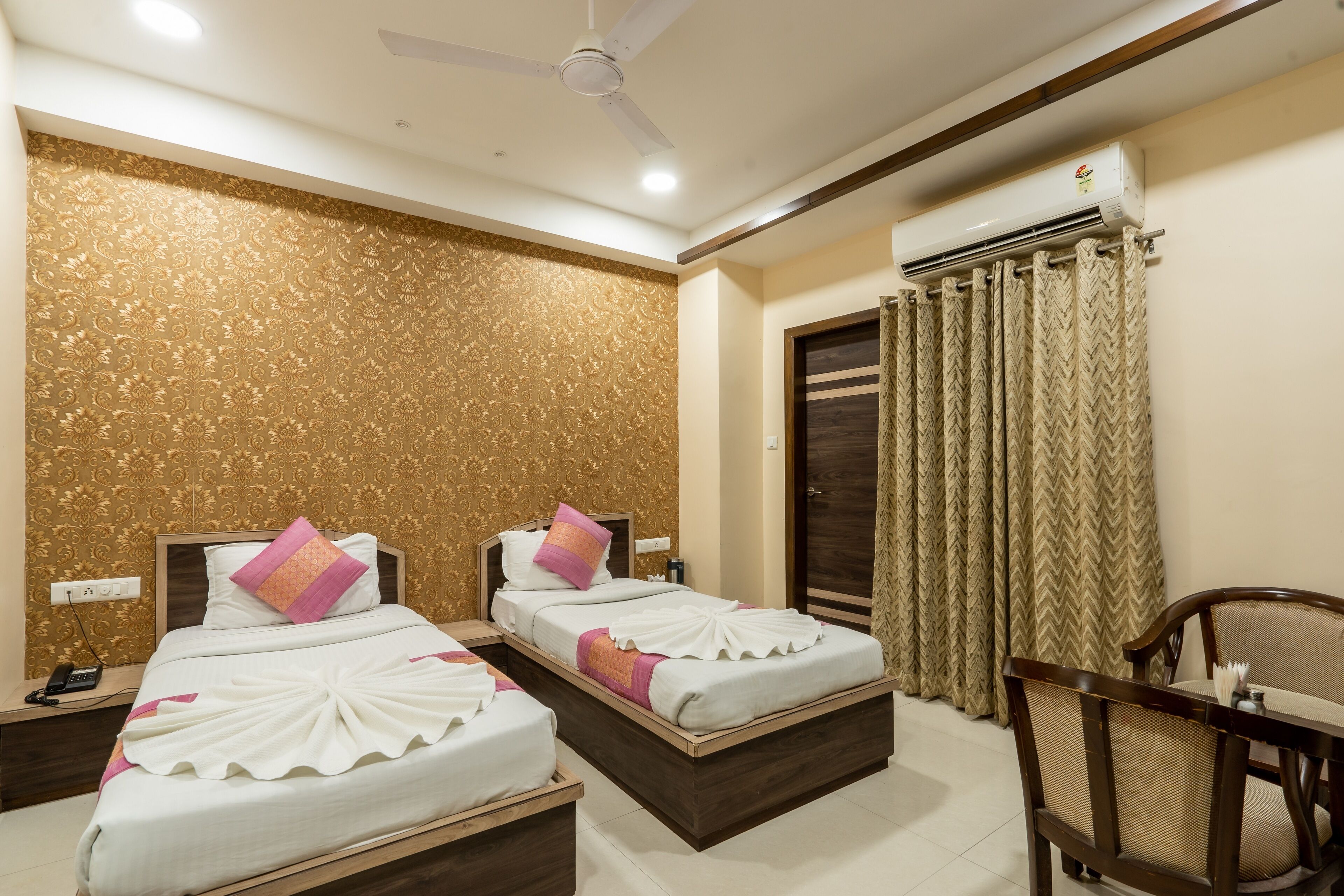 A hotel room with two beds, a wooden headboard, a ceiling fan, an air conditioning unit, a wooden table with chairs, and a patterned wall.