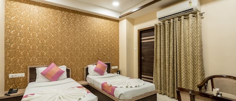 A hotel room with two beds, a wooden headboard, a ceiling fan, an air conditioning unit, a wooden table with chairs, and a patterned wall.
