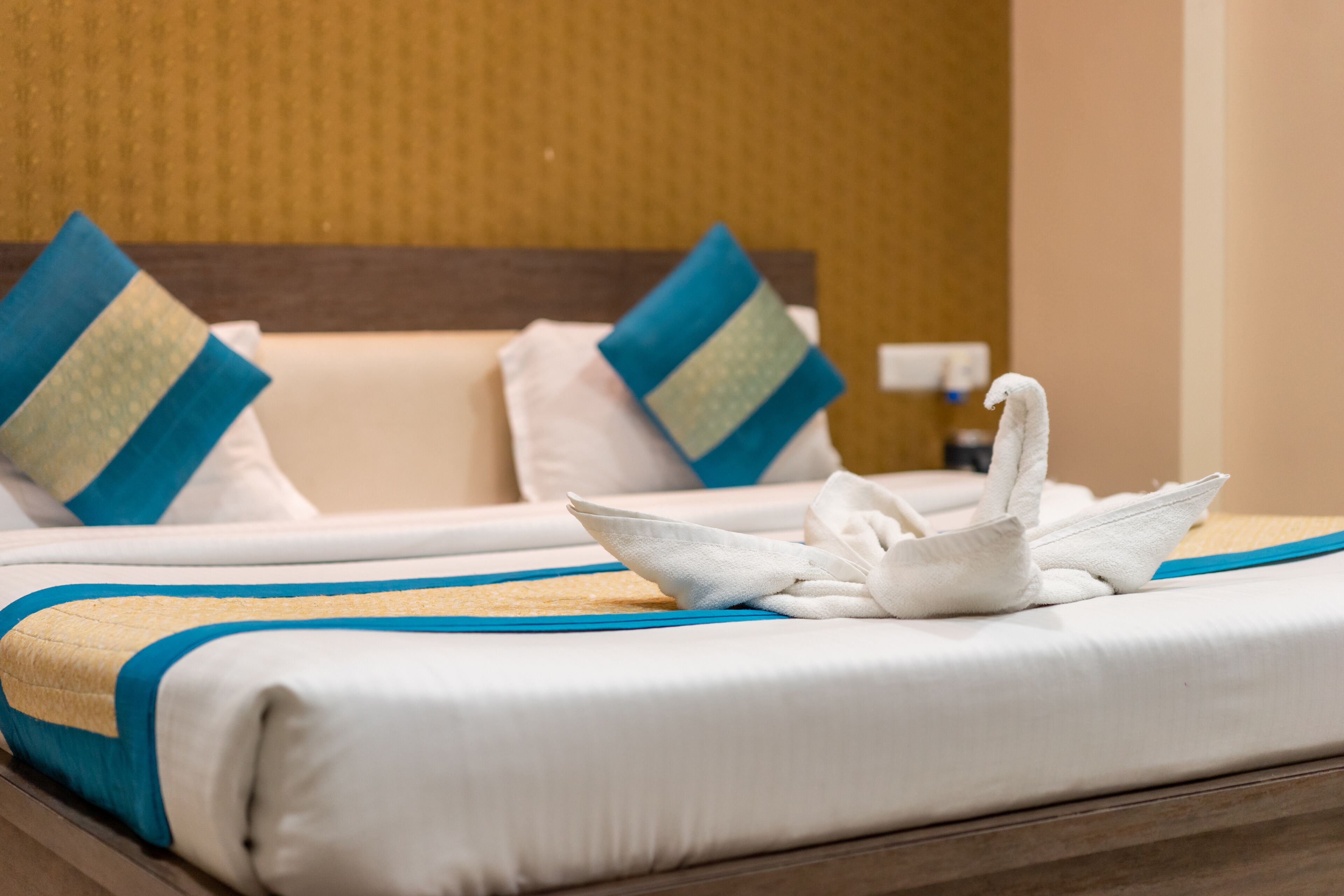 A neatly made bed with a white and blue bedspread, two pillows with blue and green patterns, and a white swan-shaped towel on the bed.