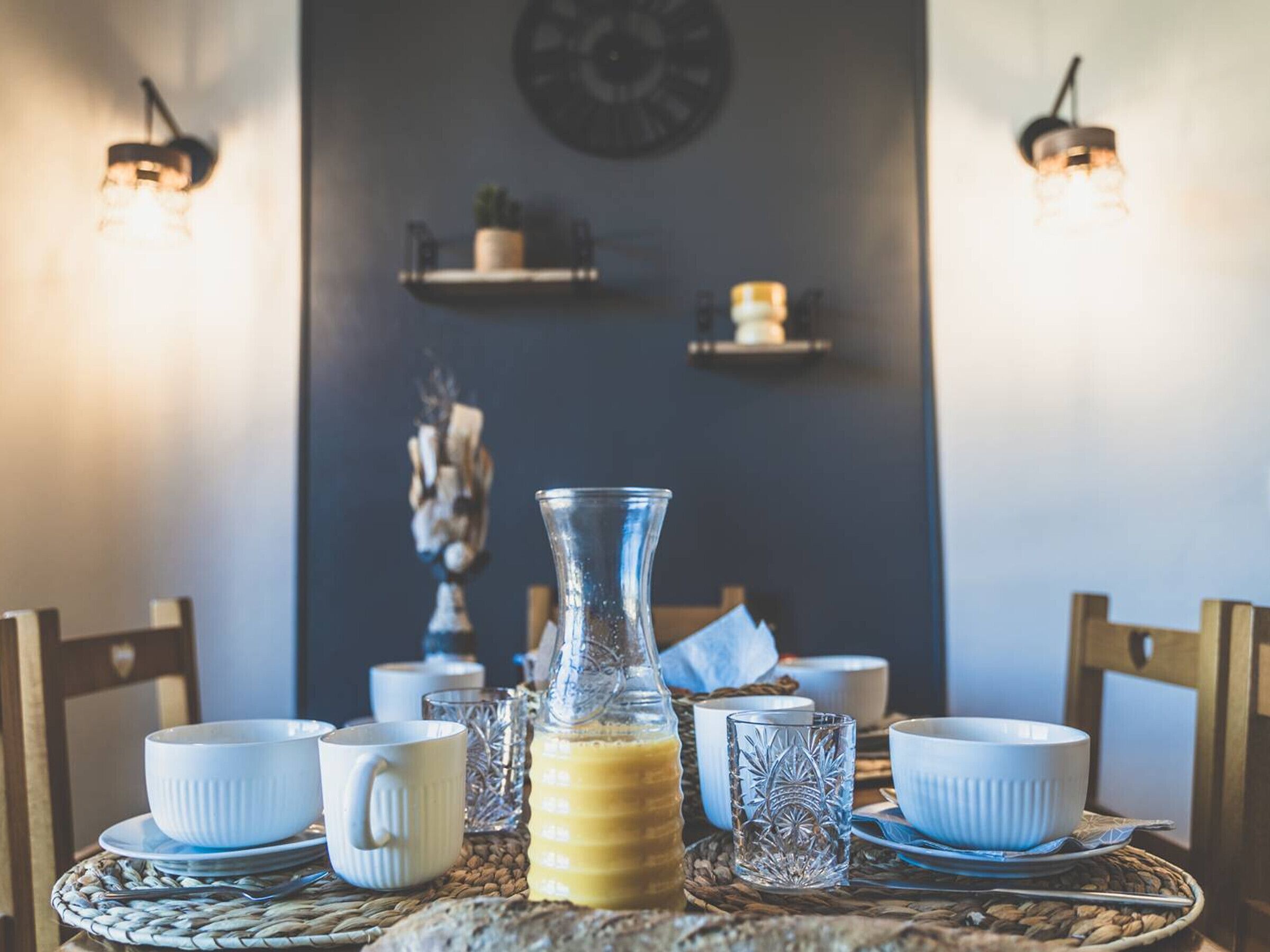 A breakfast setting with a table set for two, including a glass pitcher of orange juice, white cups, and a woven placemat.