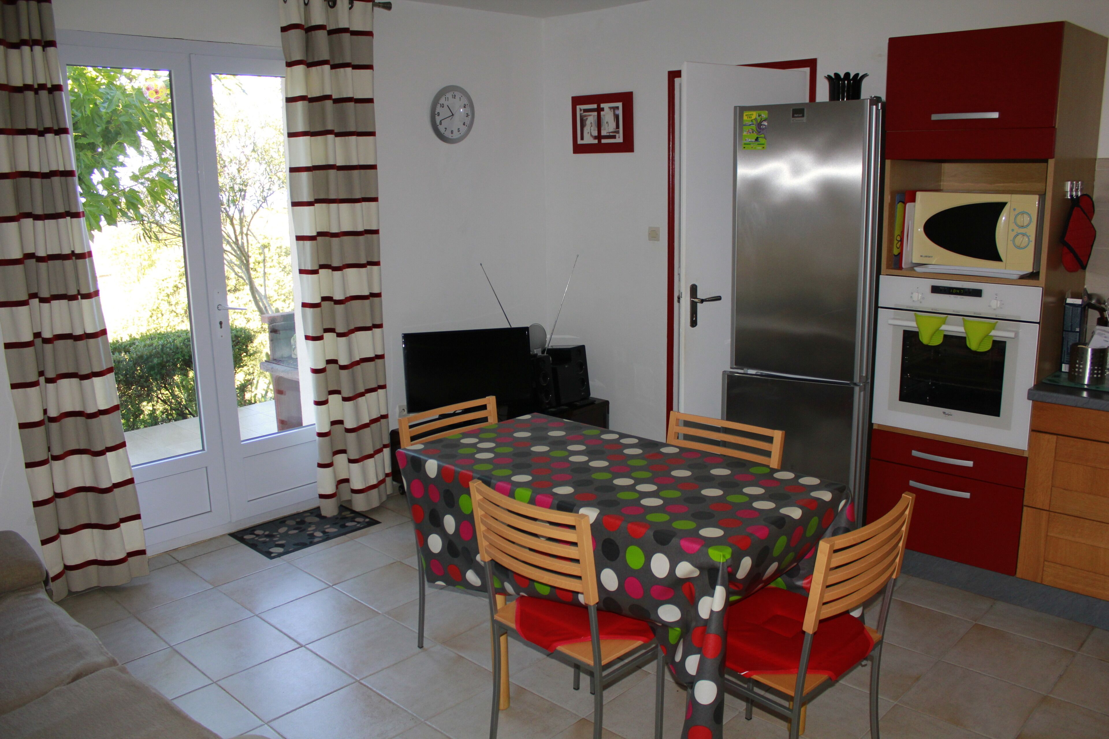 A dining area with a table covered in a polka dot tablecloth, surrounded by wooden chairs with red cushions. A refrigerator and microwave are built into the cabinetry, and a clock is visible on the wall.