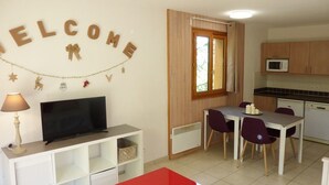Una sala de estar moderna con televisor, una mesa roja y un comedor con sillas moradas. Se observa una cocina con armarios de madera y un microondas.