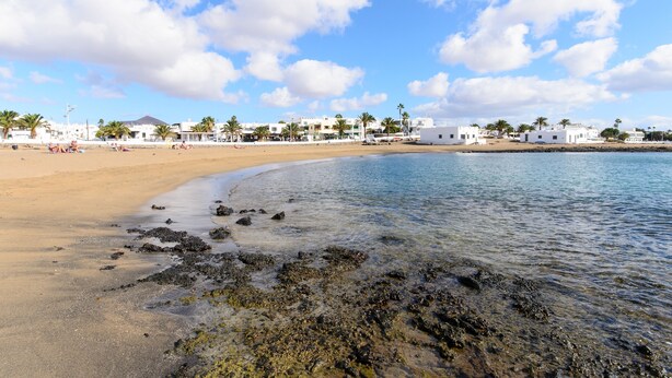 Una playa con agua cristalina, una orilla arenosa y edificios blancos al fondo.