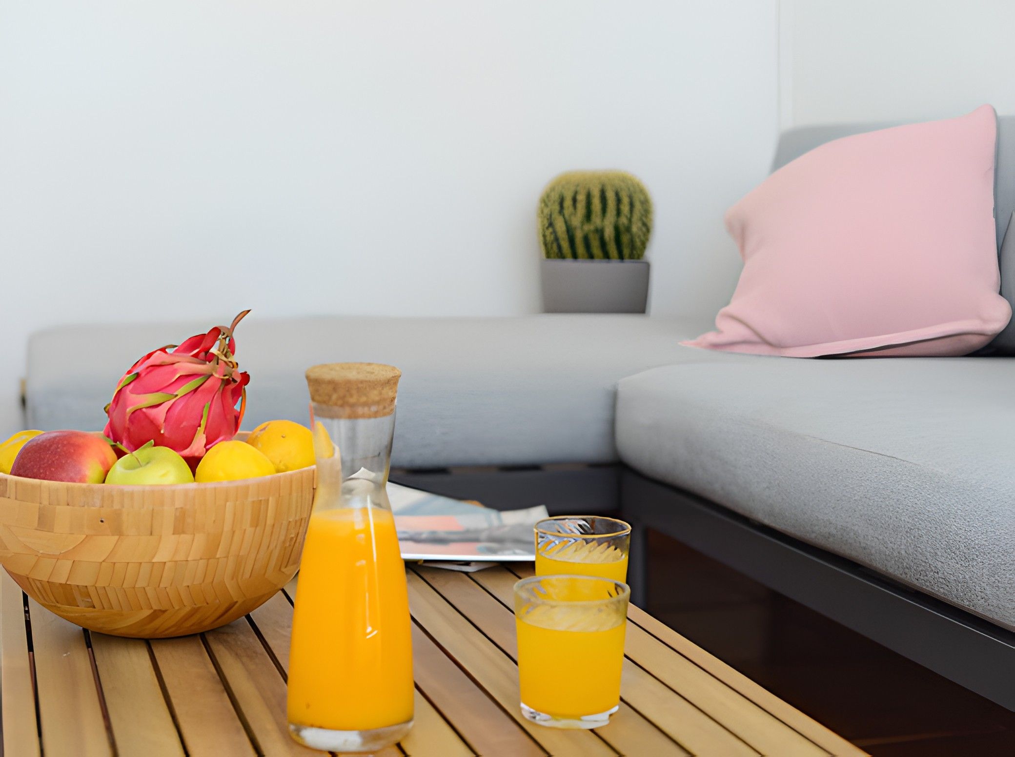 Une table en bois sur laquelle se trouvent un panier de fruits, une bouteille de jus d’orange et deux verres de jus. En arrière-plan, on aperçoit un canapé gris avec un coussin rose et une plante, un cactus.