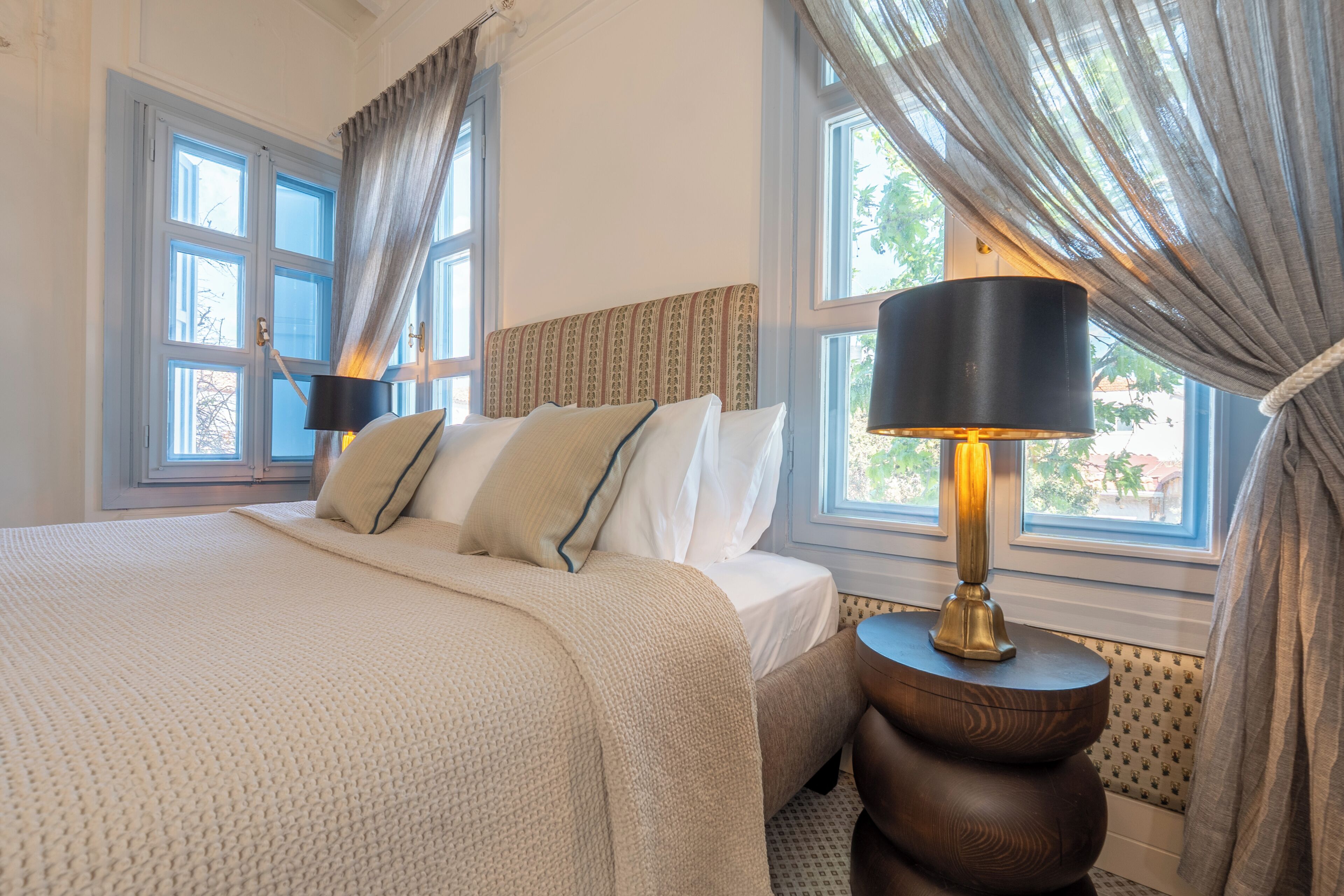 Ein Schlafzimmer mit einem großen Bett, zwei Nachttischlampen und Blick ins Freie durch Fenster.