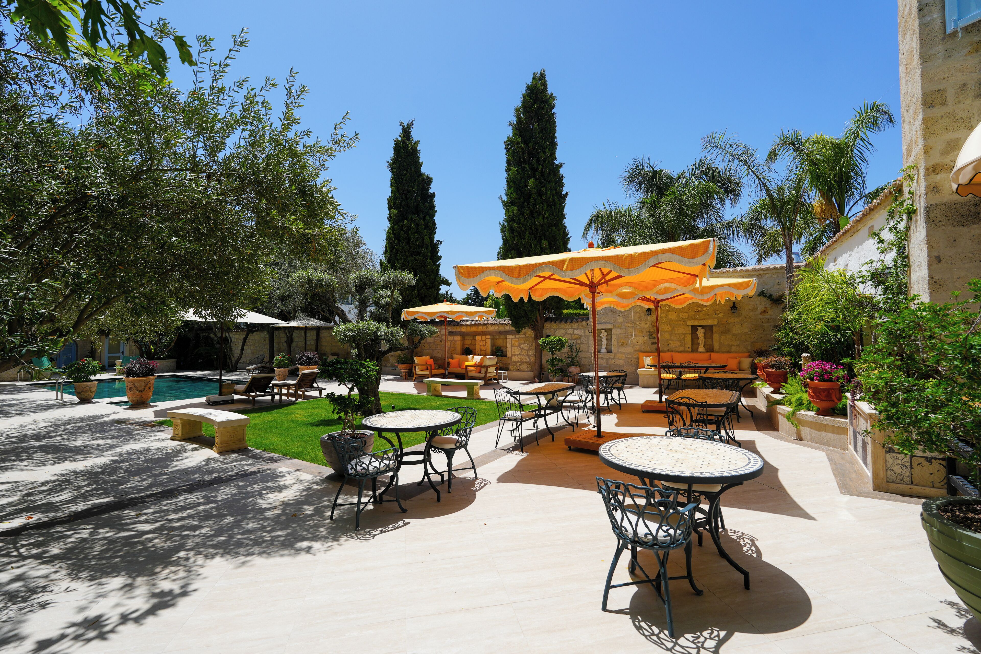 Un patio al aire libre con mesas y sillas redondas, rodeado de plantas en macetas y una piscina.