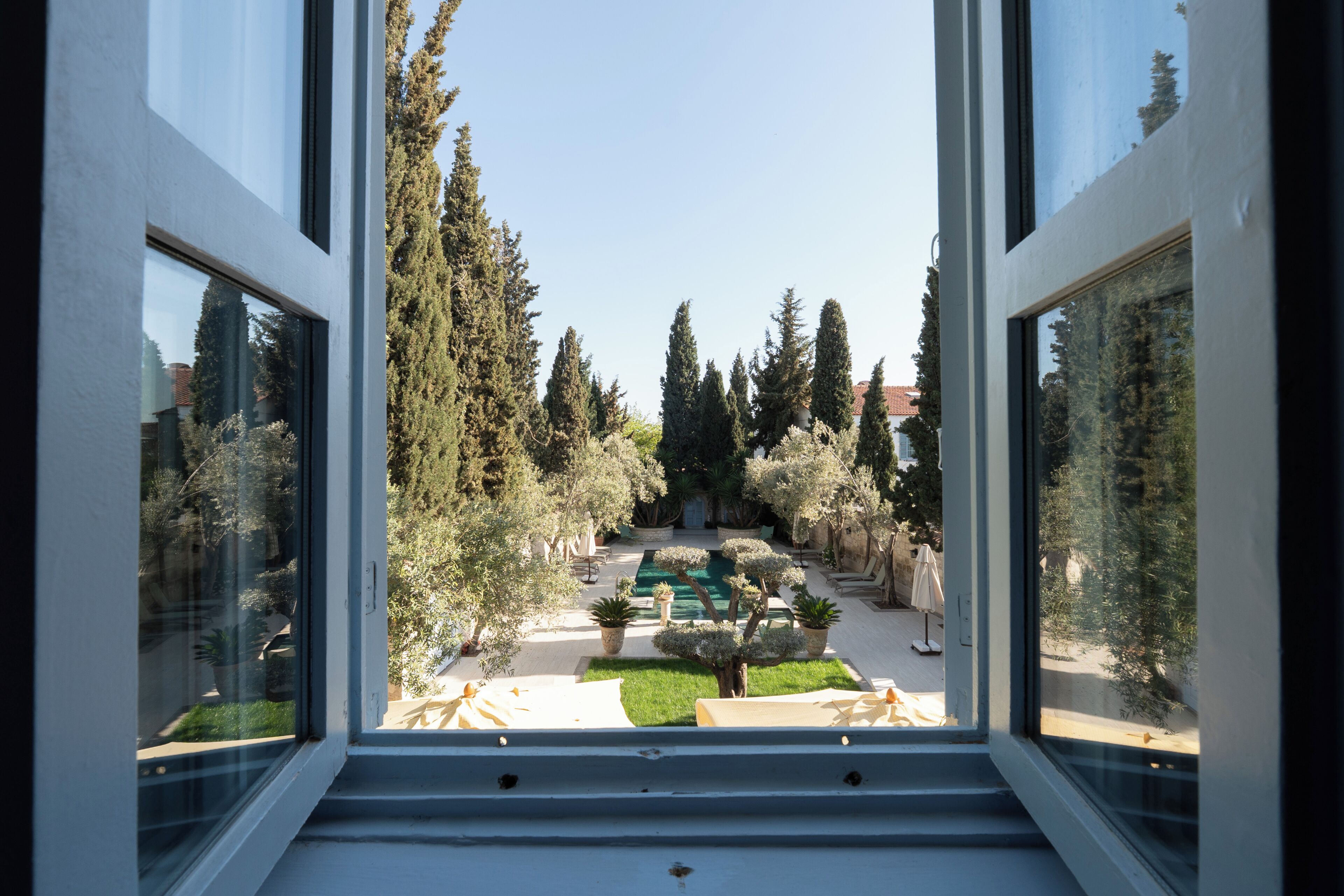 Vista desde una ventana a un patio con piscina, plantas en macetas y cielo despejado.