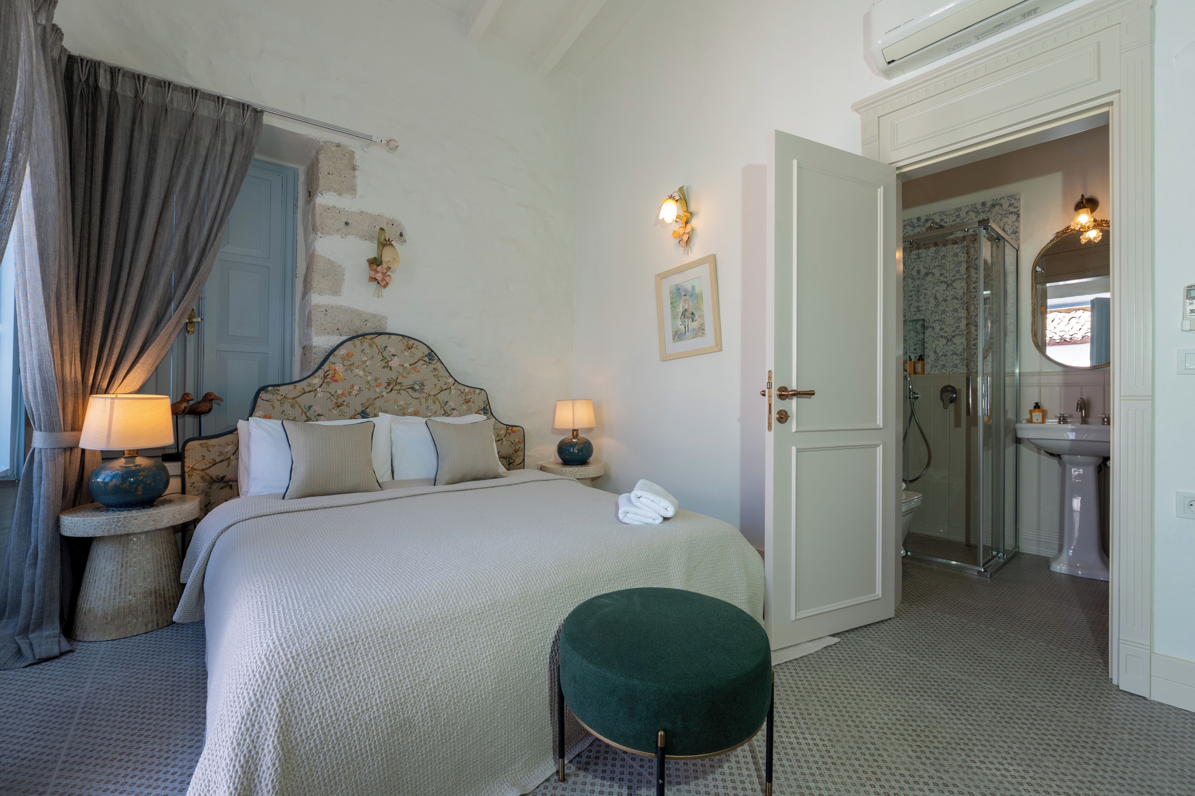 A bedroom with a bed, bedside lamps, a green ottoman, a bathroom with a sink and shower, and a door leading to another room.