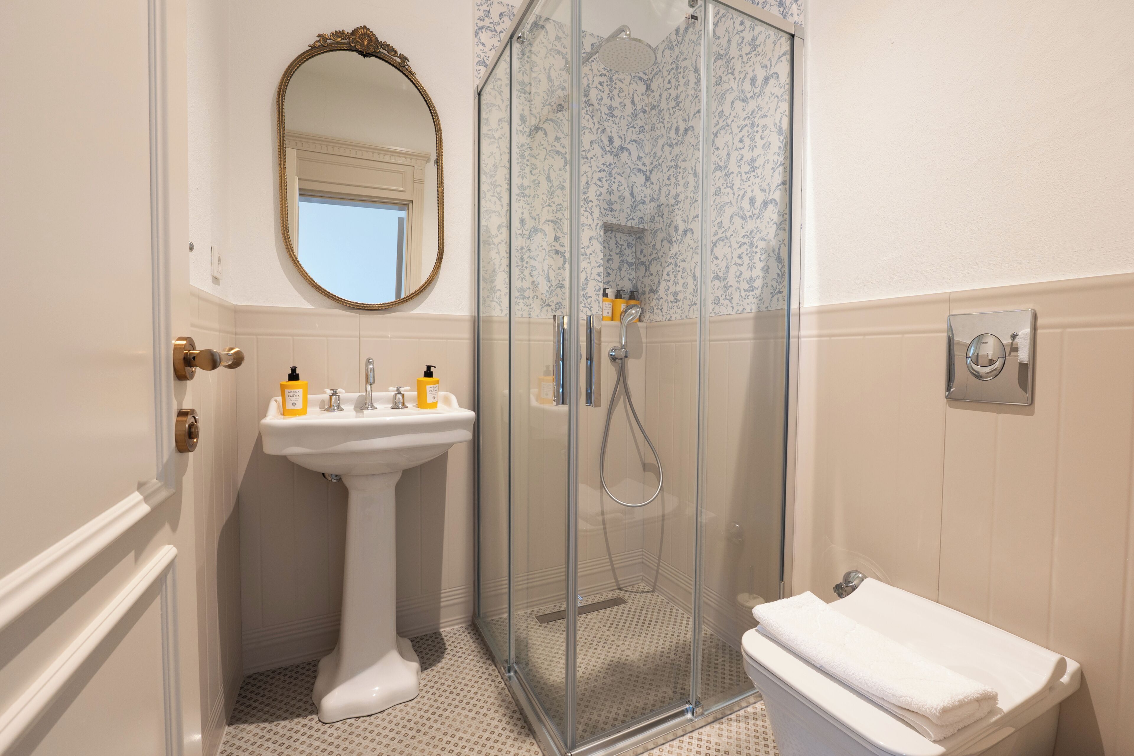 A bathroom with a pedestal sink, a glass-enclosed shower, a toilet, and a mirror with an ornate frame.