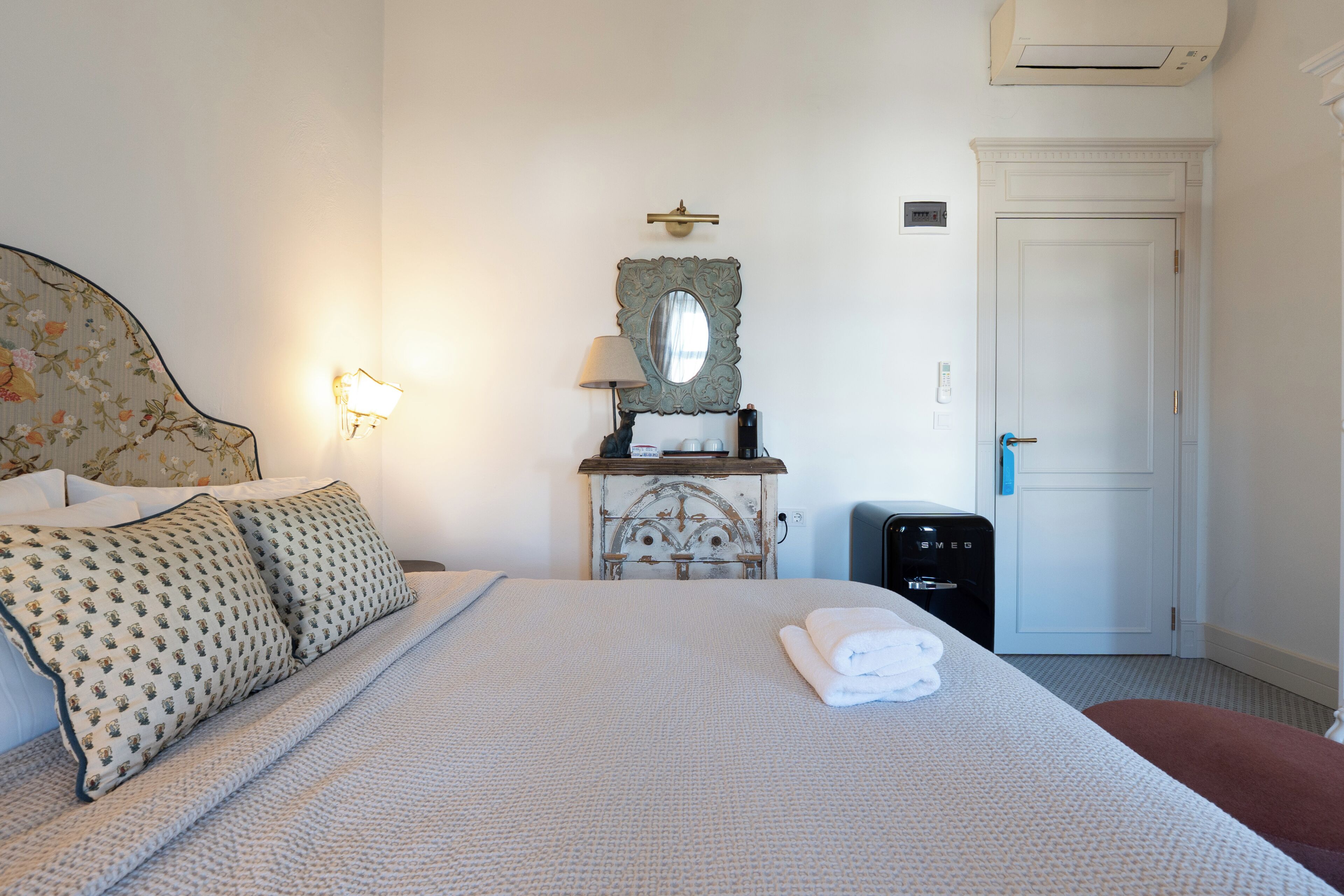 Una cama bien hecha con un almohadón estampado, una lámpara de noche, un espejo, una mesita con una lámpara y una puerta.