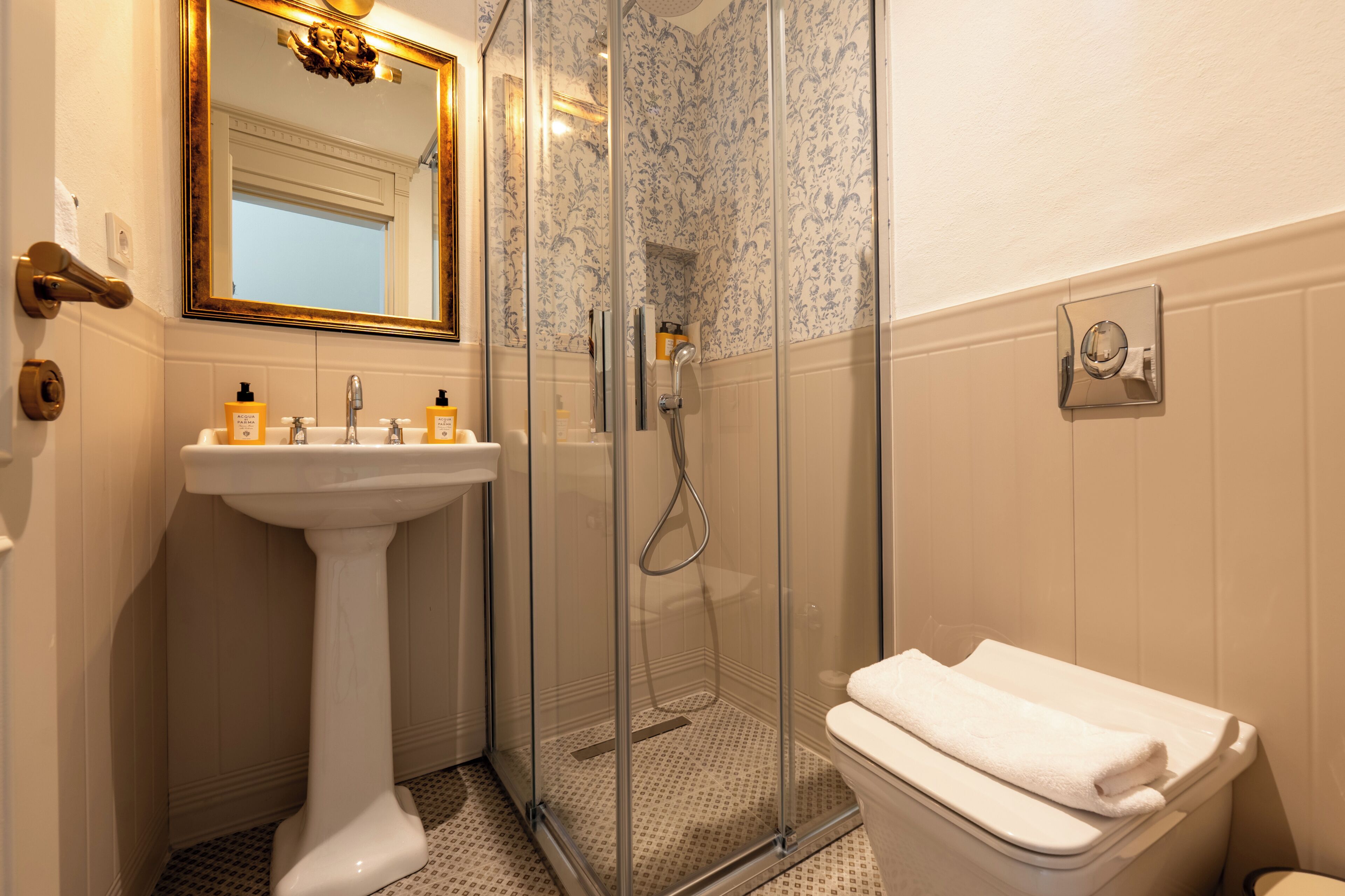 A bathroom with a pedestal sink, a glass-enclosed shower, a toilet, and a mirror with a gold frame.