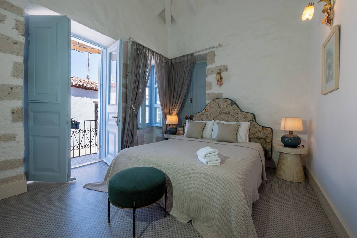 Ein Schlafzimmer mit einem Bett, zwei Nachttischlampen, einem grünen Hocker und Blick ins Freie.