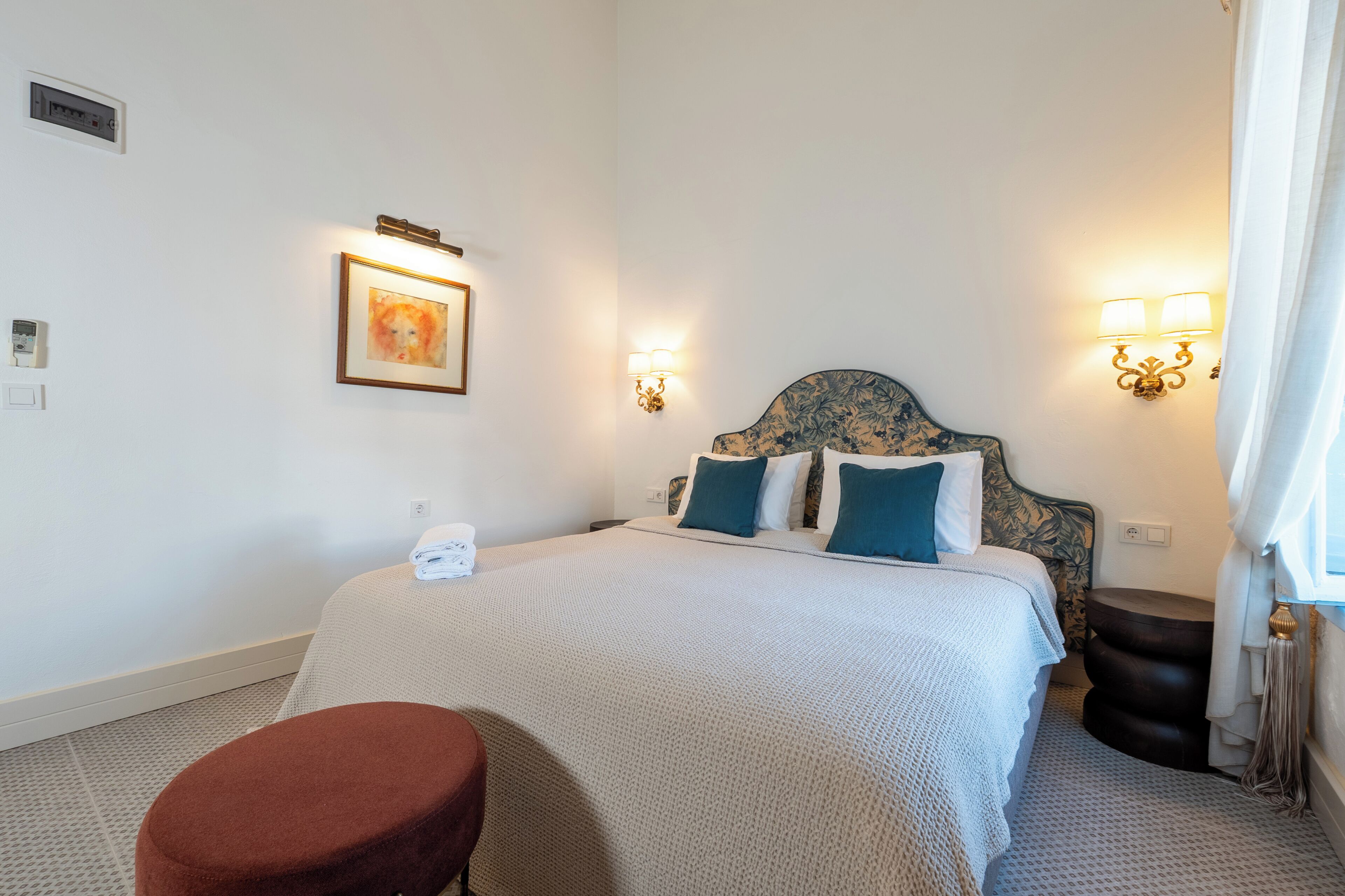 Habitación de hotel ordenada con una cama, un taburete pequeño, luces fijadas a la pared y un cuadro enmarcado.