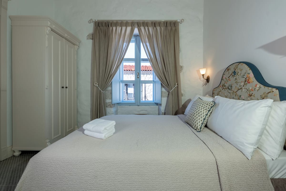 Ein ordentlich bezogenes Bett mit weißer Decke, ein Kopfteil mit Blumenmuster und ein Fenster mit Vorhängen.
