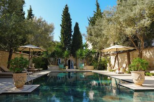Zona de piscina con sillas de descanso y sombrillas, rodeada de árboles y un edificio de piedra.