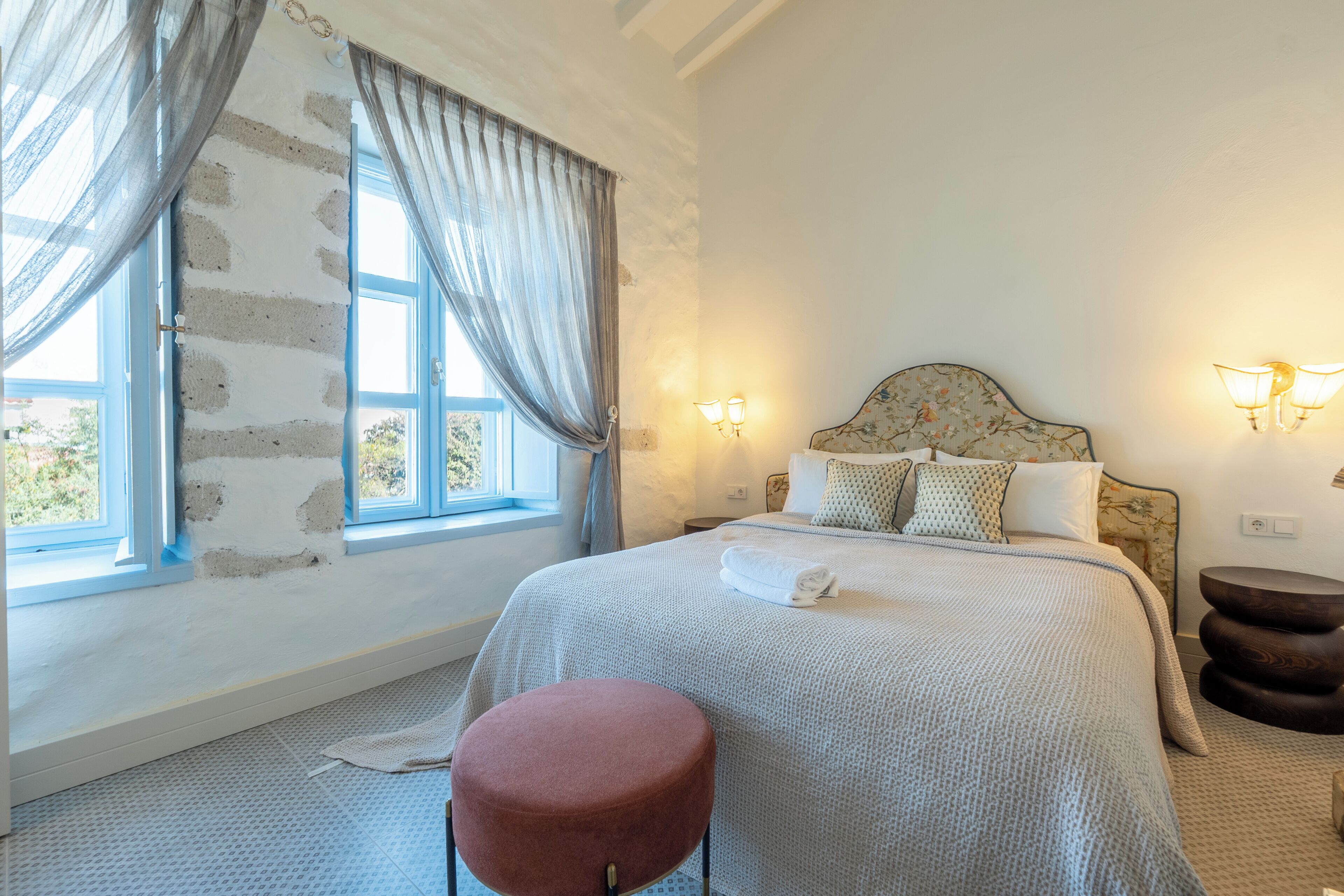 A bedroom with a bed, a window with curtains, a bedside lamp, and a small stool.