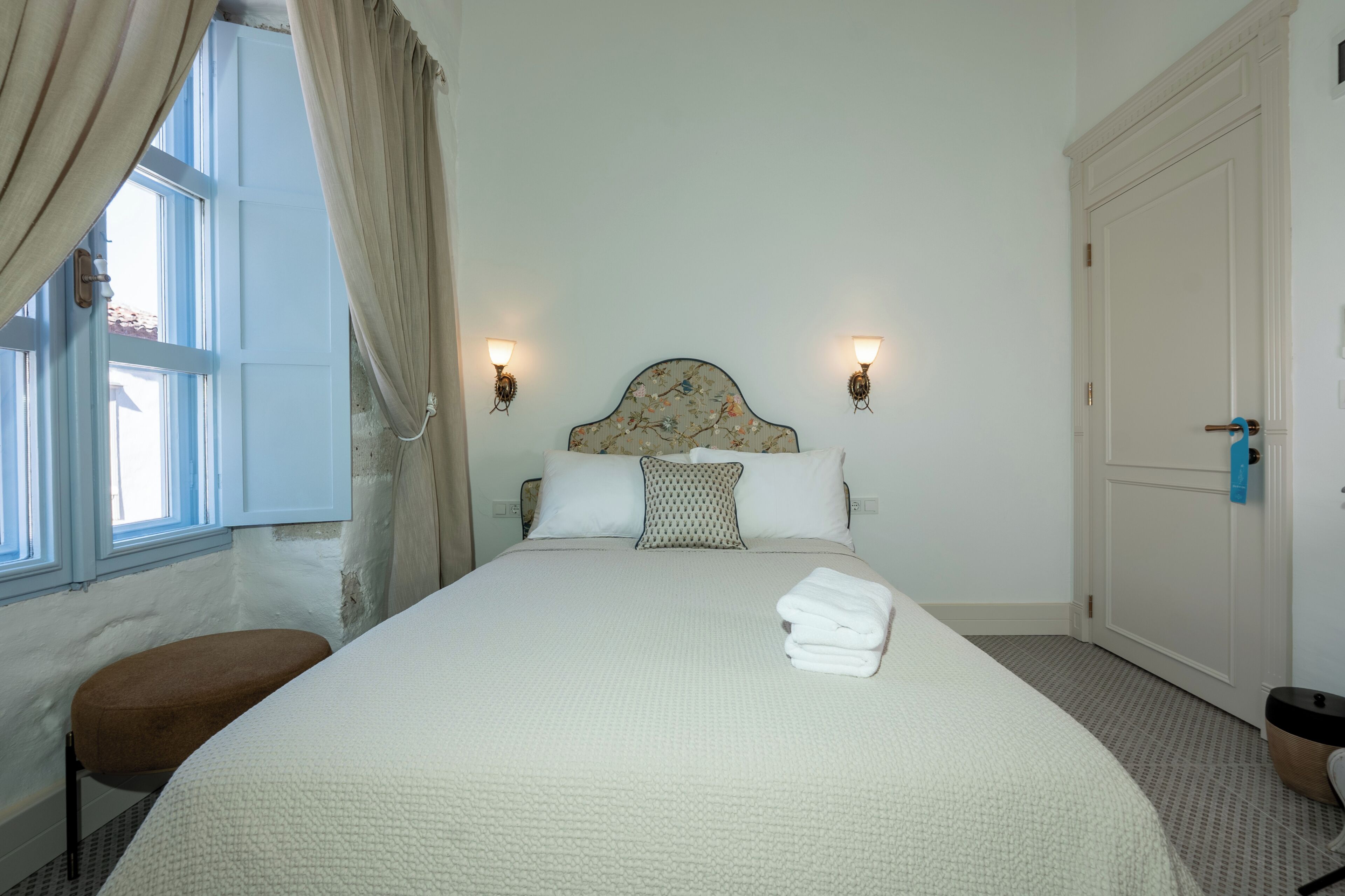 A neatly made bed with a headboard, two wall-mounted lights, a window with curtains, and a door.