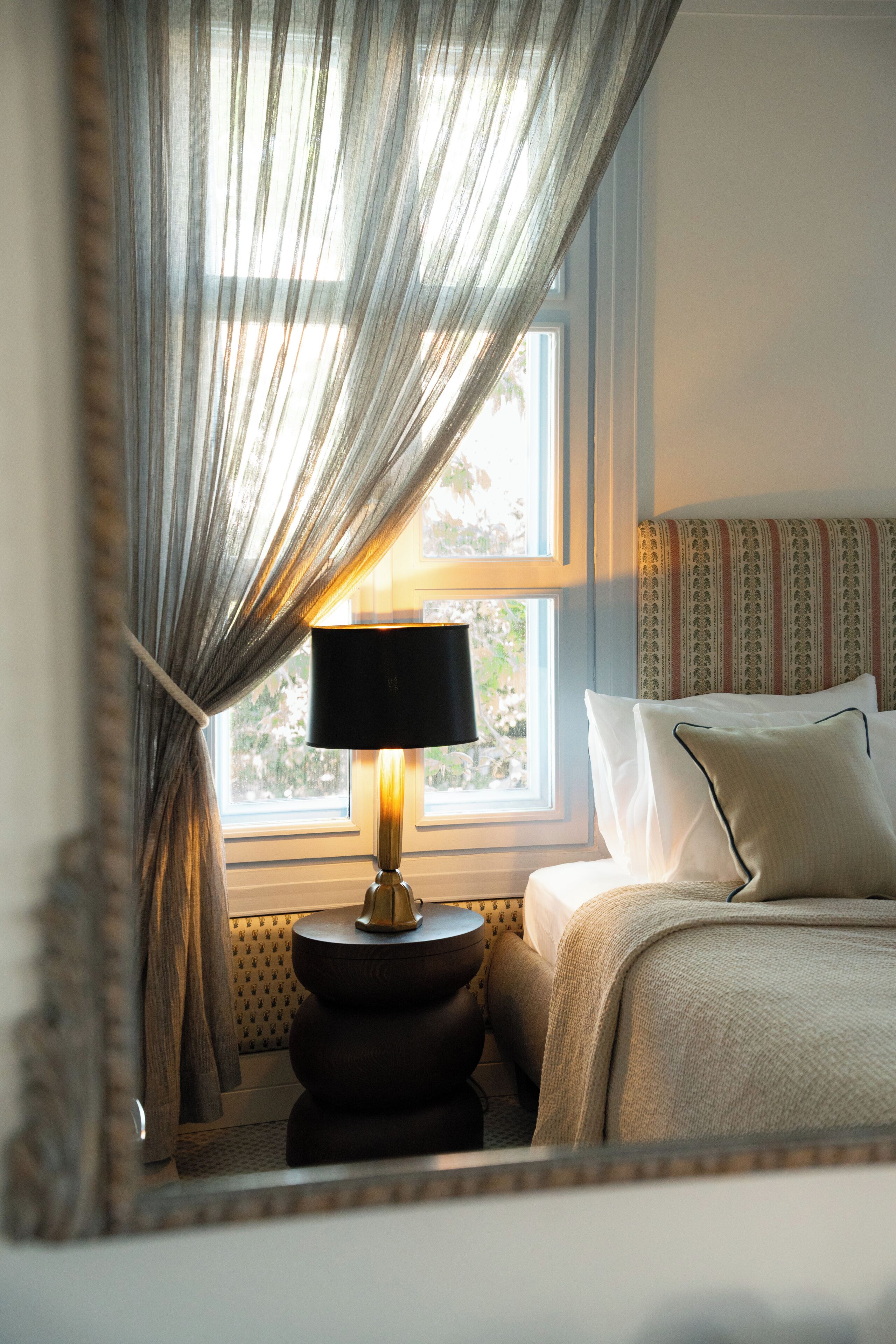A bedroom with a bed, a bedside lamp, a window with curtains, and a patterned headboard.